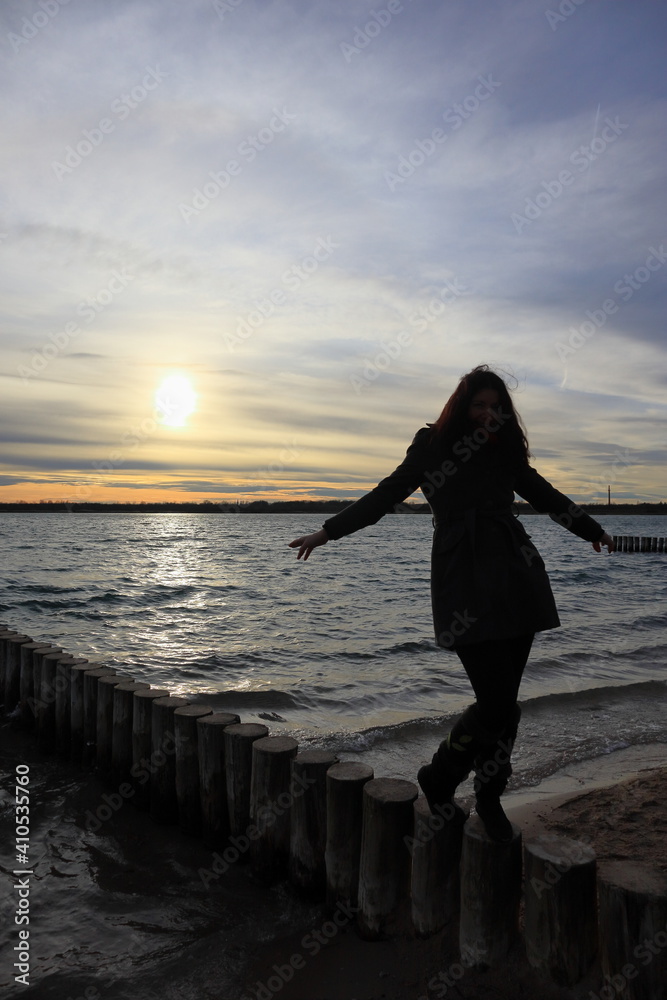 Fototapeta premium Silhouette einer jungen Frau, die bei Sonnenuntergang auf einer hölzernen Buhne am Markkleeberger See balanciert, Sonnenuntergang am Markleeberger See, bei Leipzig, Sachsen, Deutschland