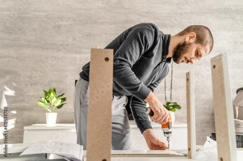 One Man with beard alone Putting Together Self Assembly Furniture at Home holding electric screwdriver looking the instructions - half length front view DIY concept real people copy space