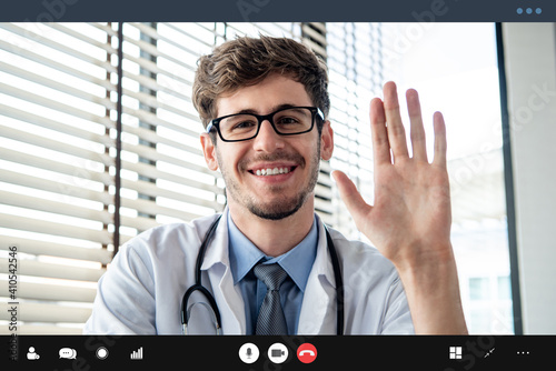 Young male doctor waving hand greeting patient online via video call, home medical consulation service concepts photo