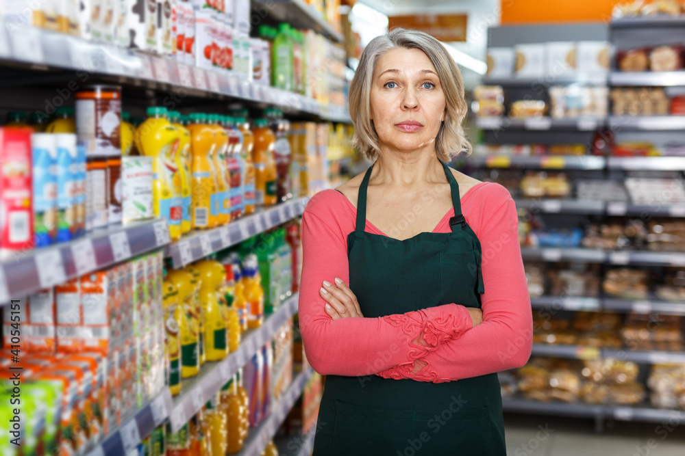 Portrait of confident mature saleswoman standing arms crossed next to shelves of food products in grocery