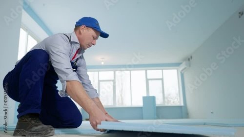 repair in new apartment, a professional man in work suit and cap on the floor lays the expanded polystyrene for thermal insulation in room photo