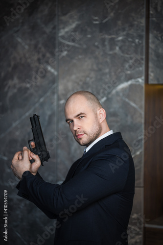 A bald man in a suit with a gun is standing against the wall in a dark room