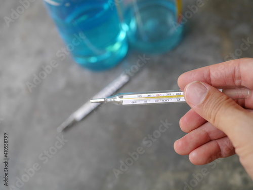 Hand held a close-up of a glass thermometer. Behind there is a picture of hand washing alcohol during the Covid-19 outbreak.