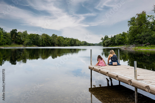 Young mother and daughter sitting on edge of a wooden deck with their backs to the camera looking in to the water, lake at Almonte, Ontario. photo