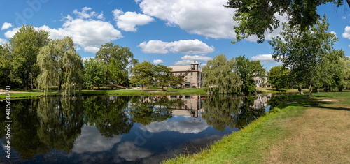 Panorama of Stewart Park, a view of the willow trees and stone house, surrounded by other trees, reflected on pond in summer. Perth, Ontario, Canada.