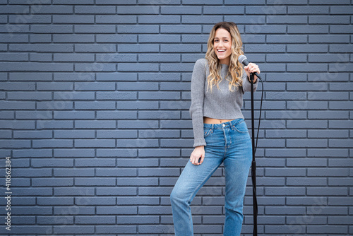 Tablou pe pânză Young Blonde Female Comedian Against Blue Grey Brick Wall with Microphone and Ex