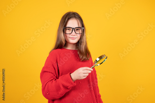 Teen girl holding magnifying glass and looking at something. Young schoolgirl in red sweater and eyeglasses on yellow background with copy space