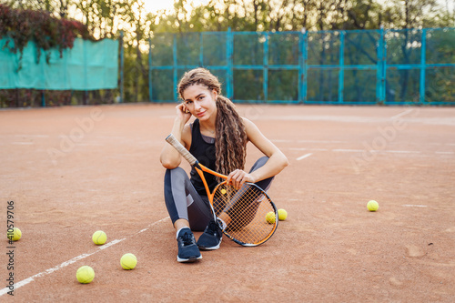 Portrait of a beautiful young woman sitting near a net tennis court among tennis balls outdoors. Sportswoman resting on a tennis court. Posing dressed in stylish sportswear © Daria