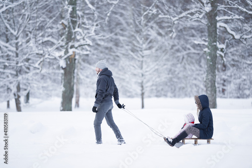 Happy young adult father pulling wooden sledge with baby girl and mother on snowy park road in deep fresh snow. Enjoying white winter day. Spending time together in weekend. Side view. © fotoduets