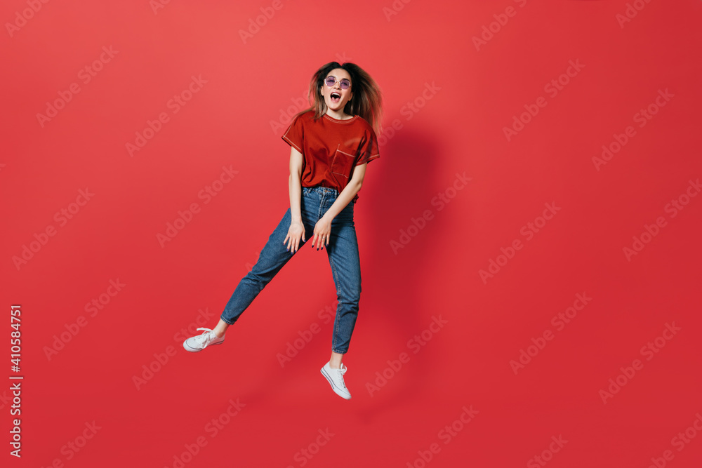 Full-length portrait of perky woman in jeans and glasses jumping on red background