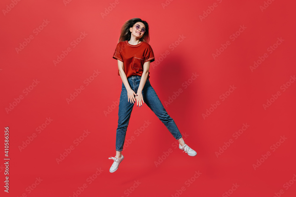 Cute girl in jeans and bright T-shirt jumping on red background