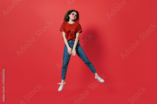 Cute girl in jeans and bright T-shirt jumping on red background