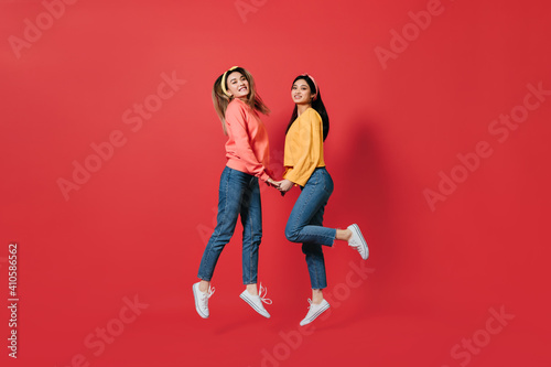 Pretty women in stylish sweatshirts and jeans jump on red background