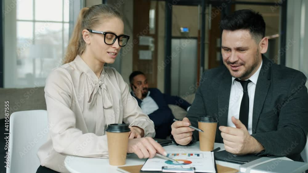 Cheerful man and woman are discussing business sitting in hotel hall at table with papers smiling conversing. People are wearing stylish formalwear and glasses. Cheerful man and woman are discussing business sitting in hotel hall at table with papers smiling conversing. People are wearing stylish formalwear and glasses.