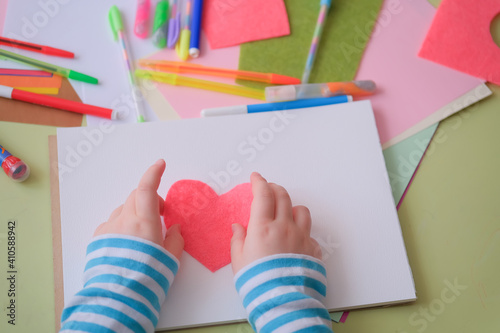   hild s boy s girl s hand holding heart cut out of felt. Children s creativity - drawings and crafts  for Valentines Day or Mother s Day or Father s Day. Top View.Colored paper  pens  scissors table