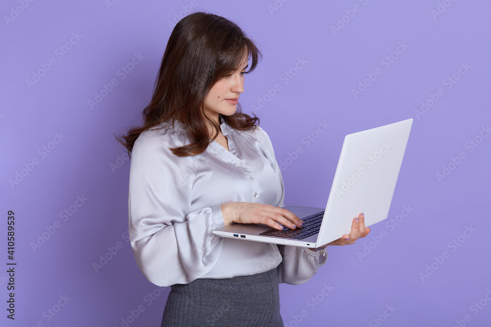 Caucasian beautiful young woman with laptop in hands posing against lilac wall, looking at device screen, being concentrated while working, wearing elegant attire.