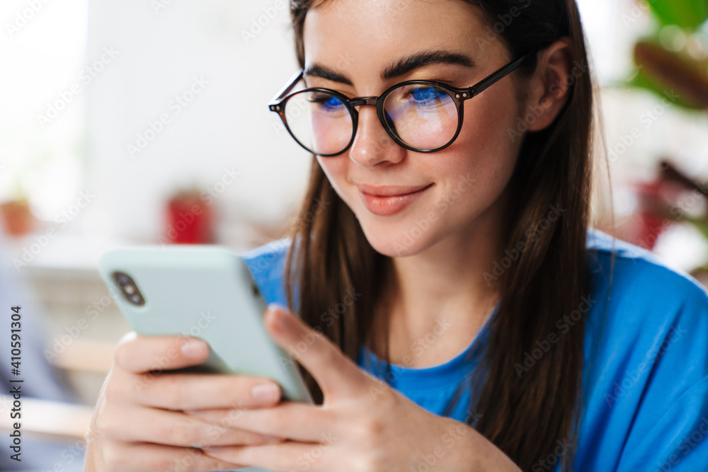 Happy brunette girl in eyeglasses using smartphone and smiling at home