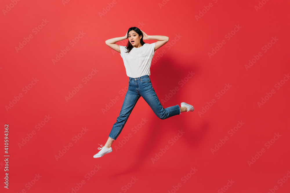 Emotional Asian woman in jeans jumping on red background