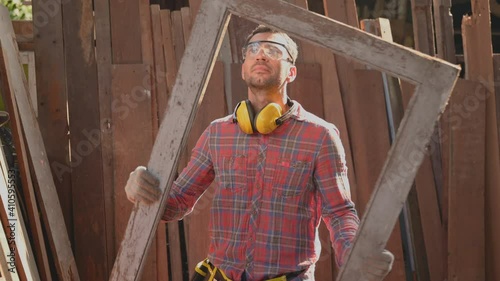 Carpenter young man wearing safety goggles standing with checking perfection of old window jamb for repair in workplace at furniture factory. Concept of small business owner and startup about woodwork
