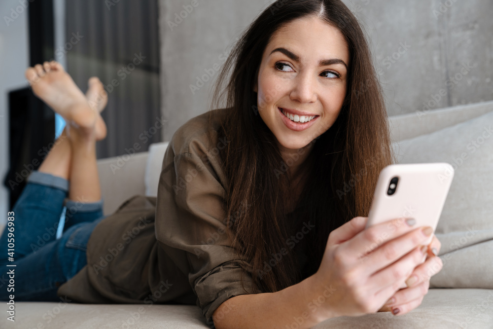 Joyful brunette nice girl smiling and using mobile phone on sofa Joyful brunette nice girl smiling and using mobile phone on sofa