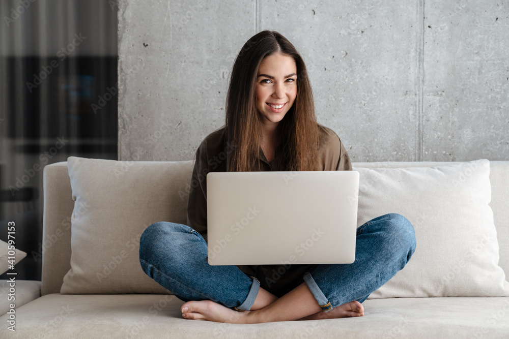 Naklejka premium Cheerful brunette nice girl smiling and using laptop on sofa