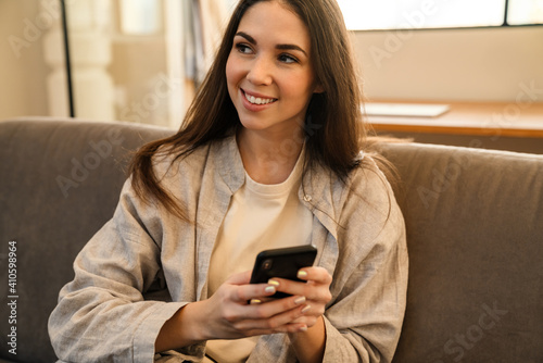 Happy young woman smiling and using smartphone while sitting on sofa
