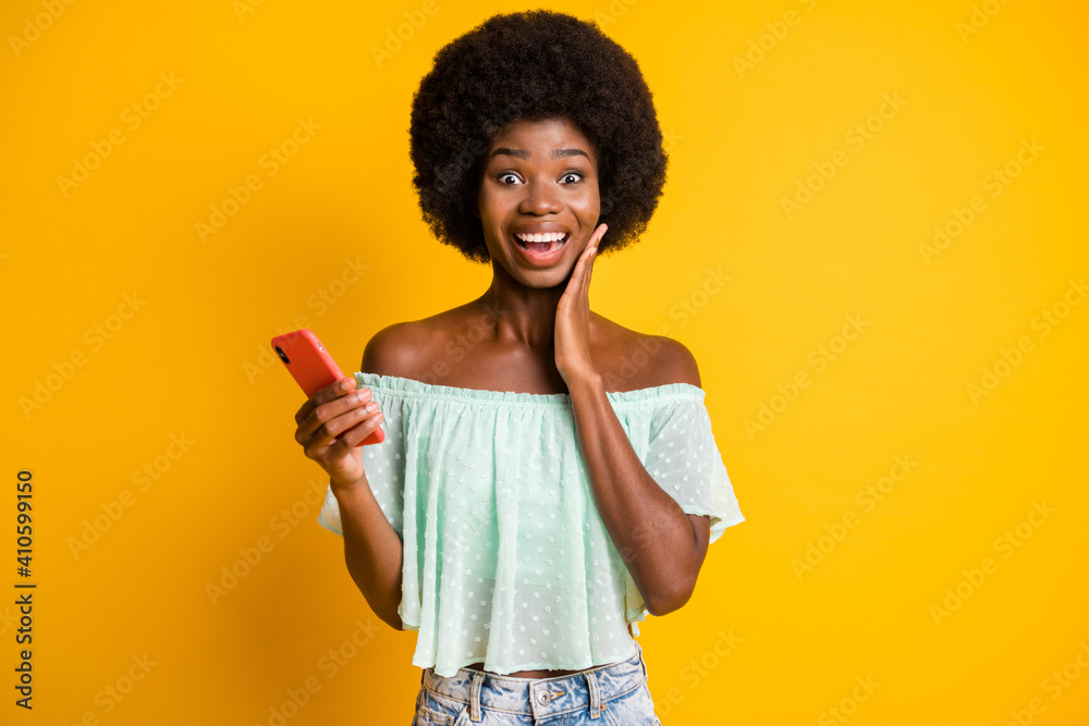 Photo portrait of shocked woman holding phone in one hand touching face cheek isolated on vivid yellow colored background Photo portrait of shocked woman holding phone in one hand touching face cheek isolated on vivid yellow colored background