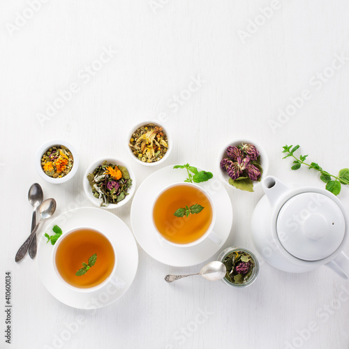 Two cups of healthy herbal tea with mint, dried rose, camomile flowers, heap of dried herbs and teapot on white wooden background