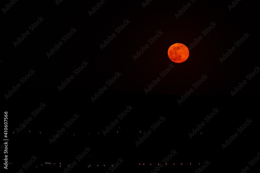 the natural satellite of the earth appearing after the horizon in bright golden colors and reflecting in the water of the lake during night summer. moonrise over the artificial lights of the village the natural satellite of the earth appearing after the horizon in bright golden colors and reflecting in the water of the lake during night summer. moonrise over the artificial lights of the village
