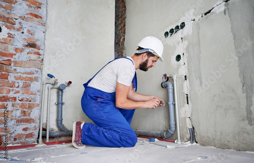 Horizontal snapshot of young plumber working with grey sewer pipes, fixing them to wall with a help of screwdriver. Side view of plumber standing on knees wearing blue uniform and white helmet
