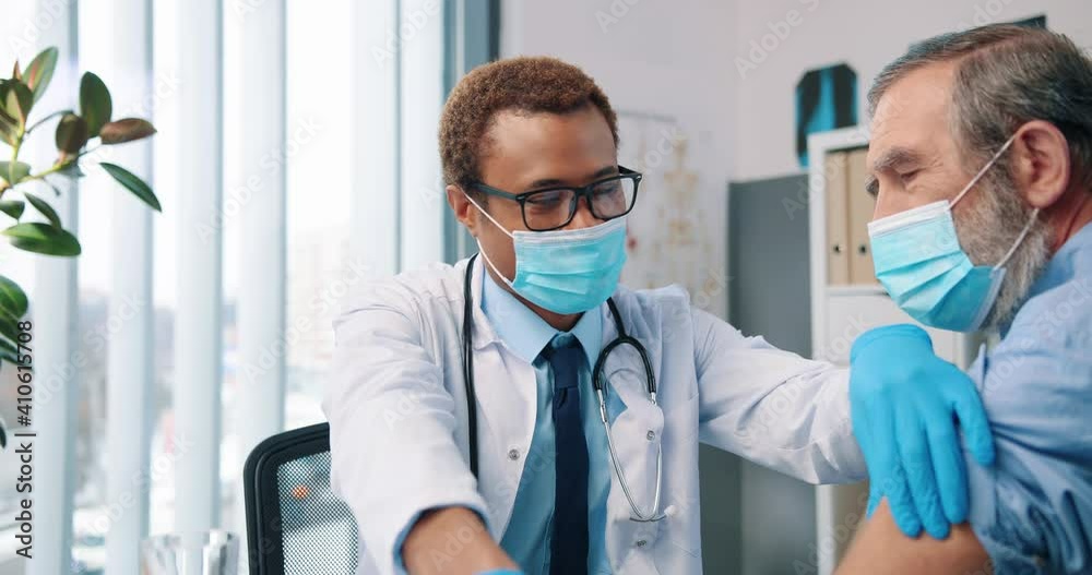Close up portrait of cheerful young African American handsome professional man doctor in white coat wearing medical mask making coronavirus vaccine injection to Caucasian old patient in clinic Close up portrait of cheerful young African American handsome professional man doctor in white coat wearing medical mask making coronavirus vaccine injection to Caucasian old patient in clinic