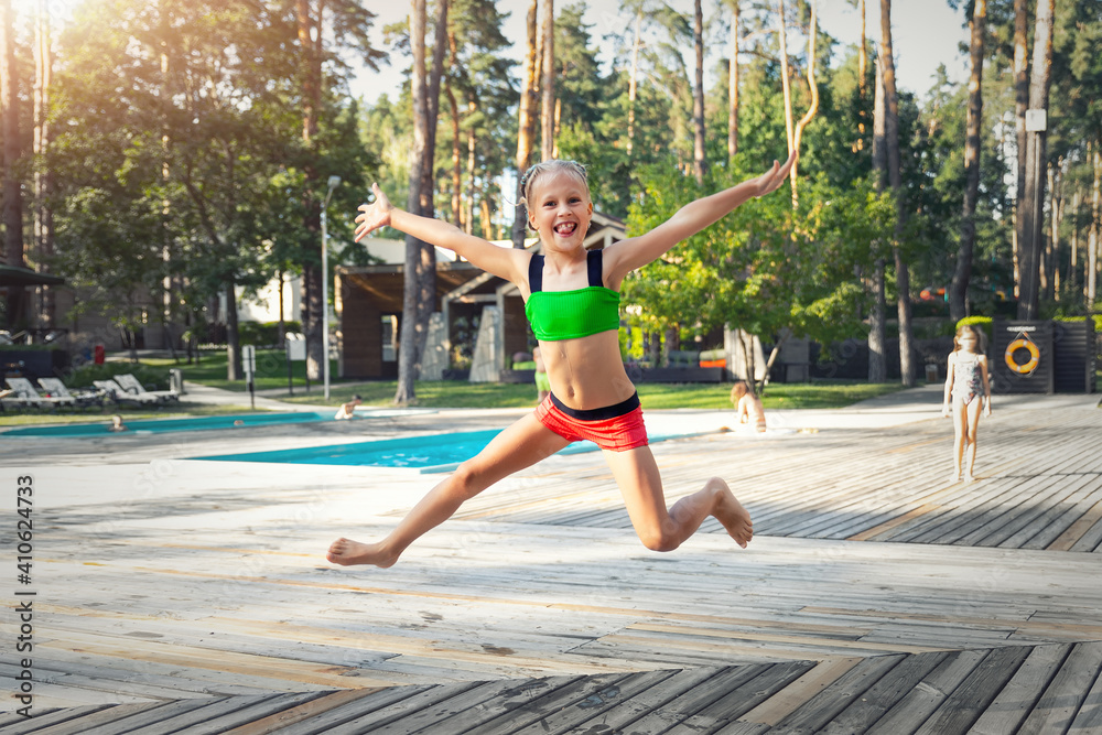 Little cute adorable caucasian blond kid girl in in swimsuit enjoy having fun jumping near pool resort area on warm sunny day. Happy child leisure and funny summer travel vacation concept. Childhood