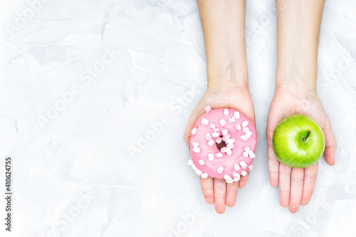 Crop view of female hands holding a fresh apple and a pink donut offering to make a choice. Healthy eating balance concept.