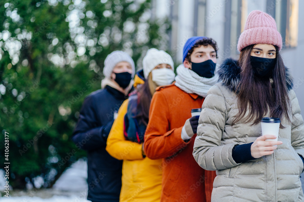 Four people friends in facial masks with coffee to go stands in row in winter time outdoor. Students in university campus standing one after another. Avoid spread of coronavirus, social distance. Four people friends in facial masks with coffee to go stands in row in winter time outdoor. Students in university campus standing one after another. Avoid spread of coronavirus, social distance.