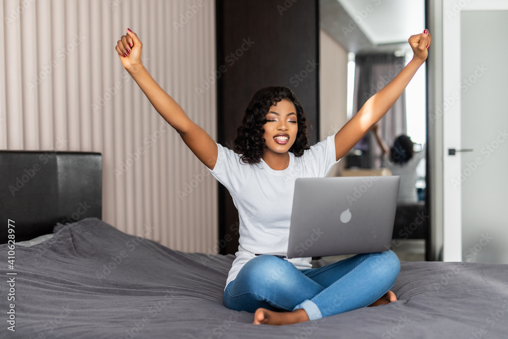 Young african american woman sitting on the bed with laptop cheering carefree and excited. Victory concept. Young african american woman sitting on the bed with laptop cheering carefree and excited. Victory concept.