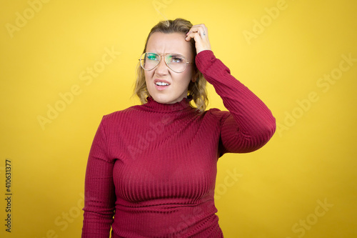 Young caucasian woman wearing casual red t-shirt over yellow background confuse and wonder about question. Uncertain with doubt, thinking with hand on head