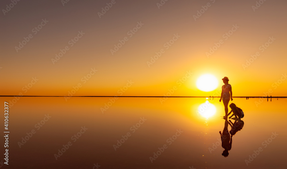 Two happy lovely sisters are walking along the mirror salt lake enjoying the evening fiery sunset