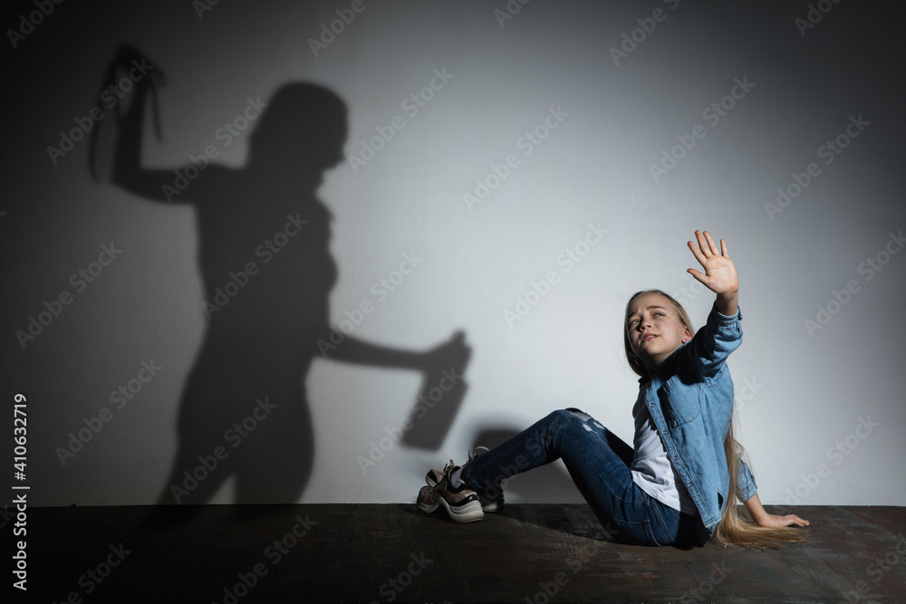 Domestic physical violence, abusing. Scared little caucasian girl, victim sitting close to white wall with shadow of angry threatening mother with alcohol addiction. Awareness of social problem.