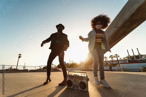 Young Afro friends wearing face mask dancing outdoor while listening to music with wireless headphones and vintage boombox
