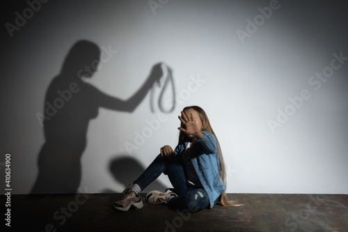 Domestic violence, abusing. Scared little caucasian girl, victim sitting close to white wall with shadow of angry mother's threatening on it. Awareness of social problem, childhood, physical violence.