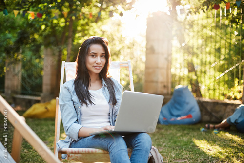Who said you cannot work outdoors. Charming smiling girl in jeans, denim jacket sit comfortable picnic chair as studying in park, feet on grass, bean chair behind, girl connect wifi and use laptop
