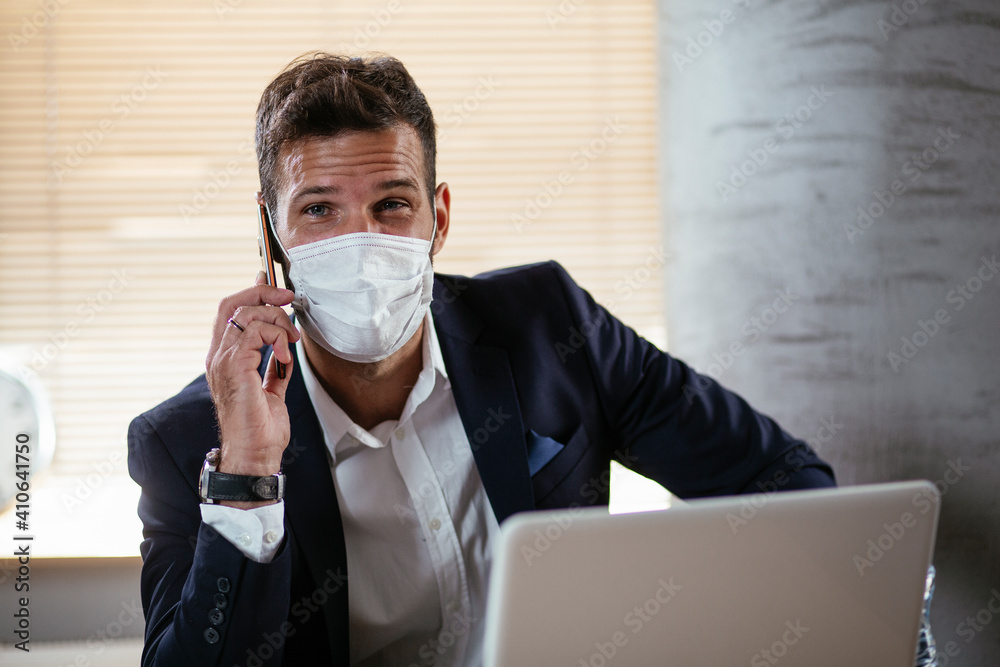 Young businessman with a mask working in his office on laptop. Businessman talking to the phone in the office. COVID-19 protection. Young businessman with a mask working in his office on laptop. Businessman talking to the phone in the office. COVID-19 protection.