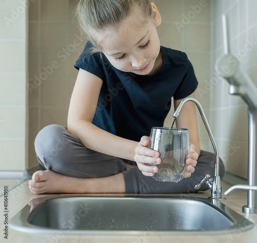 Little girl drinking from water tap or faucet in kitchen. Pouring fresh drink. Healthy lifestyle. Water quality check concept. World water monitoring day. Environmental  pollution problem