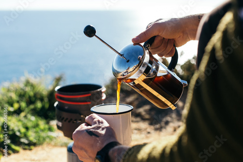 Hipster millennial man with hand tattoos prepare specialty coffee in french press, use camping stove and enamel cup. Good filter coffee during camping trip photo