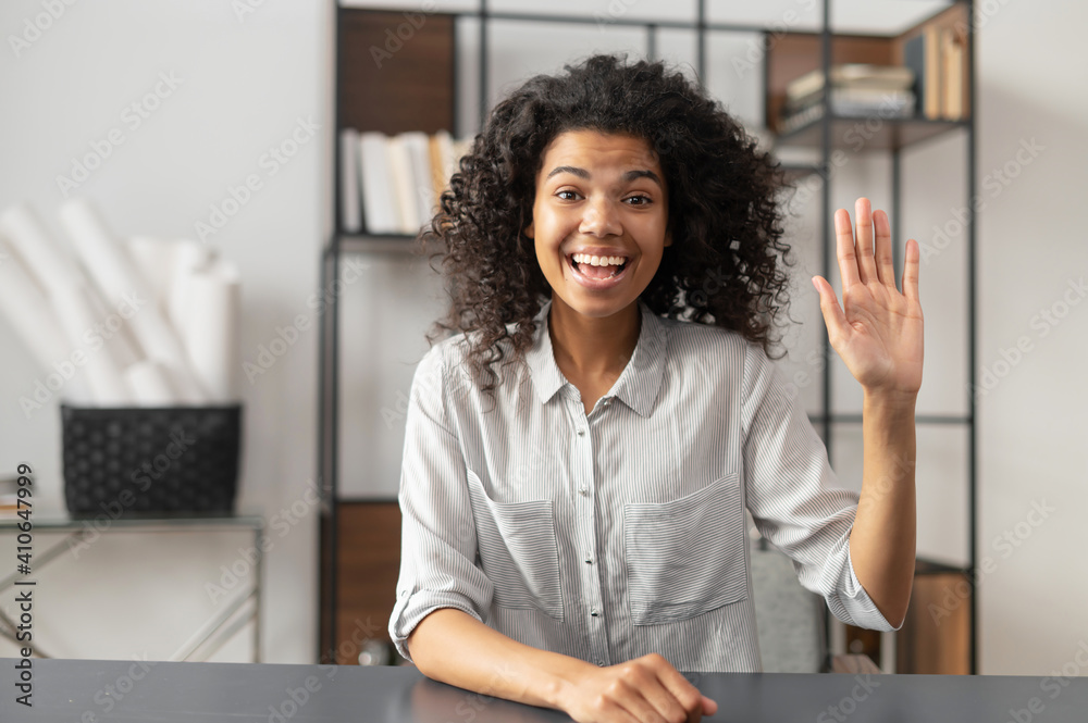 Young and energetic African-American female fresh graduate is ready for an online interview on a video call, sitting at the desk and looking and waving at the camera, saying hello. Job hunting concept Young and energetic African-American female fresh graduate is ready for an online interview on a video call, sitting at the desk and looking and waving at the camera, saying hello. Job hunting concept