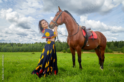 Beautiful gypsy girl with a horse in field with green glass in summer day and blue sky and white clouds background. Model in ethnic dress posing with farm animal