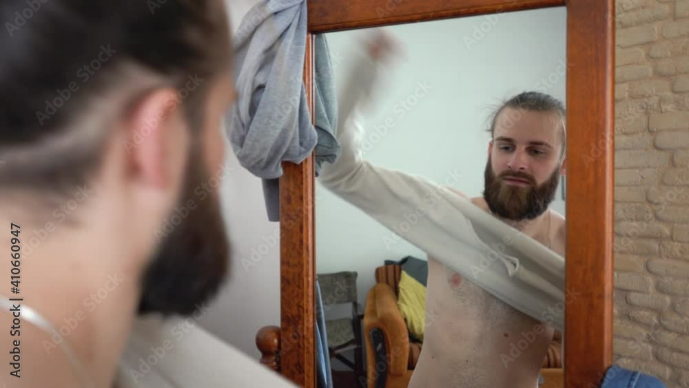 Attractive Caucasian beard man with long hair is looking into the mirror Putting together an outfit with new long sleeve T-shirt Attractive Caucasian beard man with long hair is looking into the mirror Putting together an outfit with new long sleeve T-shirt