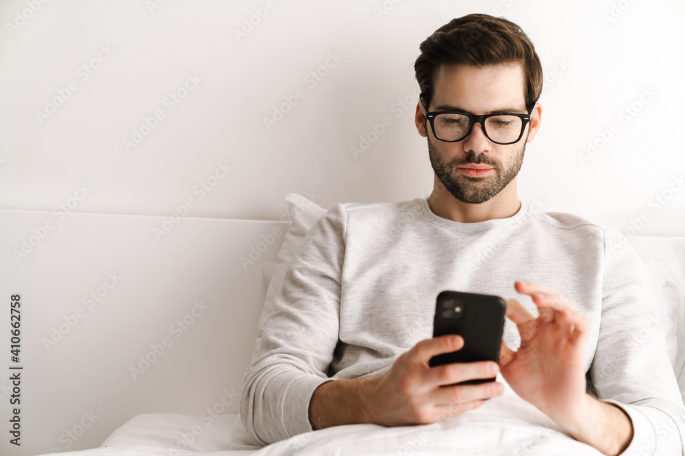 Serious young man in eyeglasses using mobile phone while resting in bed Serious young man in eyeglasses using mobile phone while resting in bed
