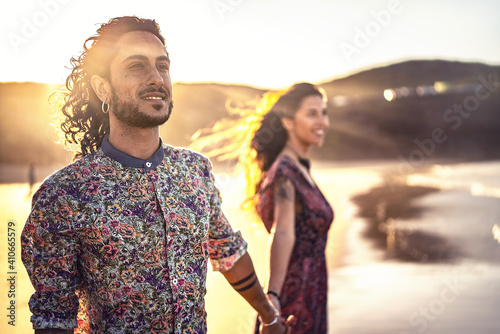 Attractive italian couple holding hands and looking together at the same direction. Romantic date at sunset on the beach .Real people emotions and love concept. Focus on the man.