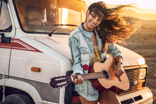 Hipster beautiful woman playing guitar with big smile on her face. Nomadic and camper lifestyle concept. Warm sunset light.